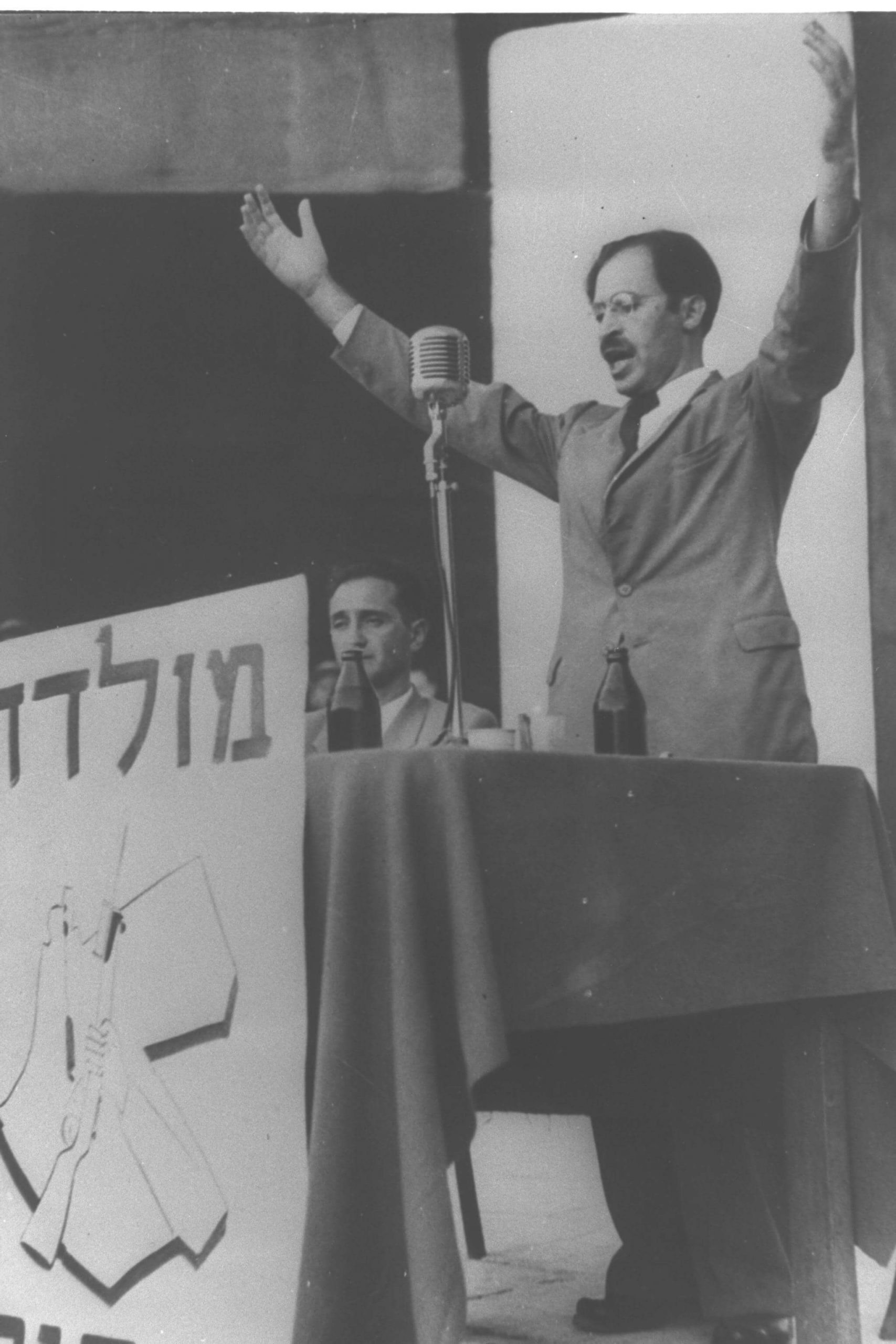 בגין נואם לאחר יציאתו מהמחתרת, ב-14 באוגוסט 1948, בתל אביב. יושב לידו חיים לנדאו. מלפניו הכתובת "מולדת וחרות" וסמל אצ"ל. © ויקיפדיה